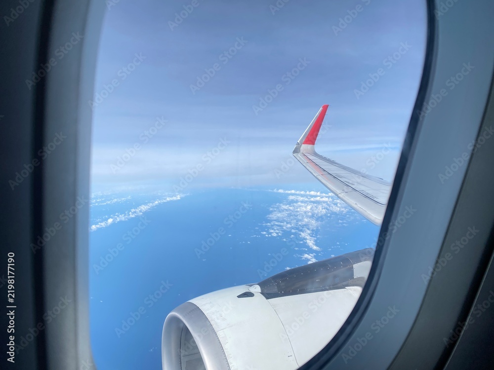 Obraz premium View from an airplane window looking out at the red and white wing tip and the engine, flying over blue ocean and white clouds under a clear blue sky