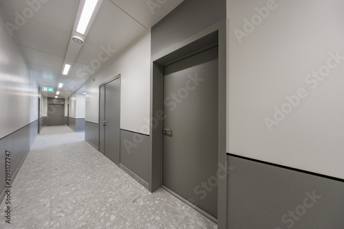 Clean modern corridor with multiple gray doors, two-tone walls, terrazzo flooring, linear ceiling lights, and an illuminated emergency exit sign. Copy space