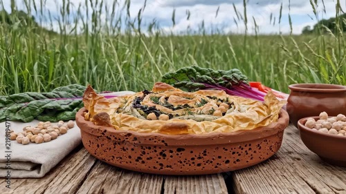 Spiral filo pie packed with leafy greens and chickpeas in a porous clay dish, framed by fresh chard, linen and meadow grass for a wholesome countryside lunch beneath soft open sky and field calm