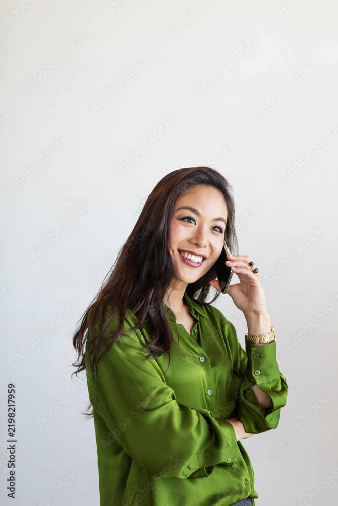 Obraz premium Smiling Asian woman in green blouse talking on cellphone, standing against white background.