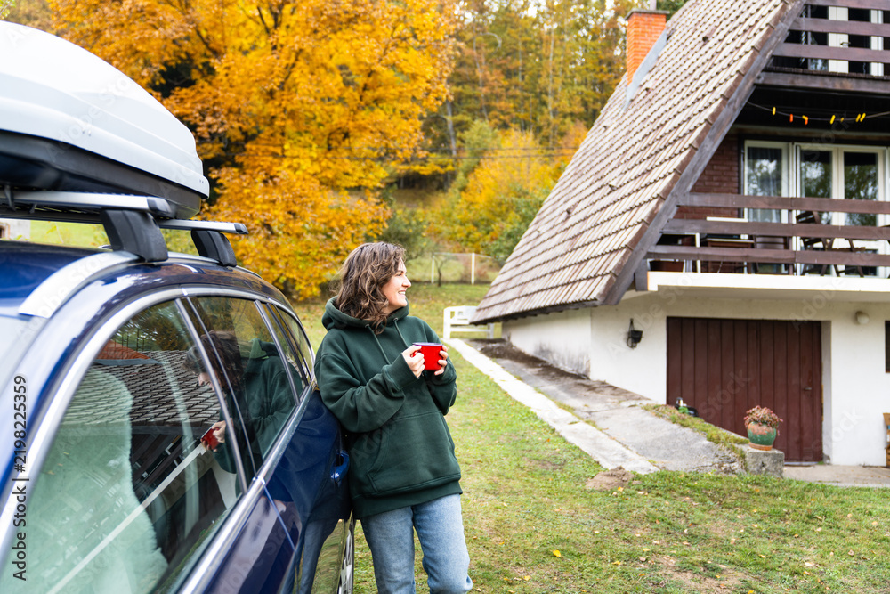 Obraz premium A woman leaning against a blue car with a red mug in her hand in front of a wooden A-frame house surrounded by colorful autumn forest