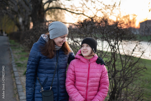 A mother and daughter walk along a riverside path at sunset, smiling and sharing warm, affectionate moment. Communication between teenage daughter and mother during a walk