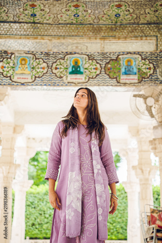 Portrait of a young Indian woman in traditional dress standing at an ancient temple in India, with carved stone pillars softly blurred in the background.