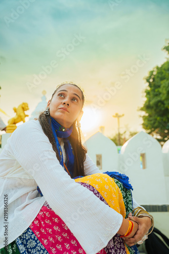 Portrait of a young Indian woman in traditional dress standing at an ancient temple in India, with carved stone pillars softly blurred in the background.