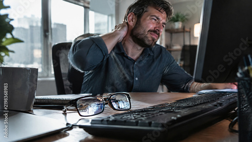 Stressed mature man rubbing his neck while working at desk in modern office