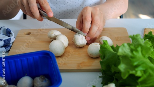 Person prepares raw champignon mushrooms on wooden board with metal kitchen knife, creating potential kitchen injury risk as bare fingers stabilize vegetable dangerously near sharp steel blade.