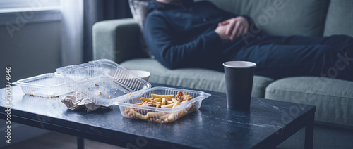 Fast food takeout meal on living room coffee table with french fries and disposable cup near man resting on couch