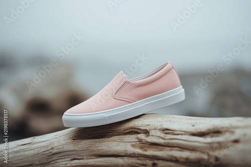 Elegant women's slip-on sneaker in soft pink balanced on a smooth piece of driftwood with a coastal background