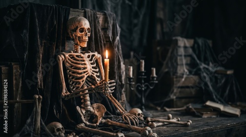 Eerie human skeleton holding a burning candle in a dark gothic room with skulls, old books and black drapery