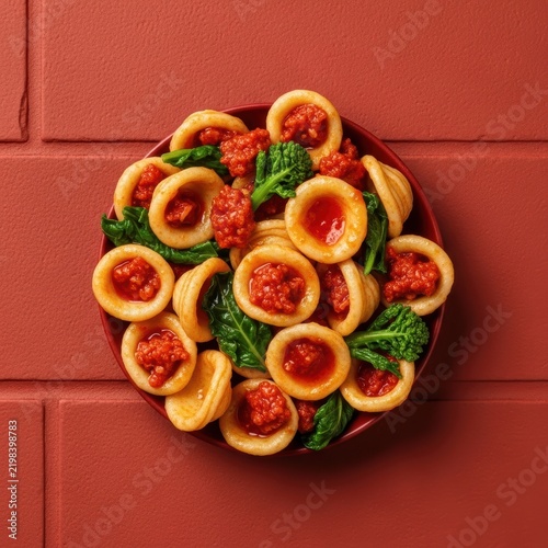 Fresh italian orecchiette pasta with savory meat sauce and green spinach leaves on a rustic red table