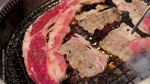 Footage of a person uses metal tongs to turn thin beef slices sizzling on a tabletop Korean barbecue grill during an interactive restaurant dining experience.
