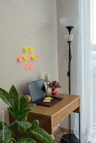 Cozy home office corner with laptop, sticky notes, pen, floor lamp and green houseplants beside bright window. Concept of remote work, task planning, online study, productivity and work life balance.