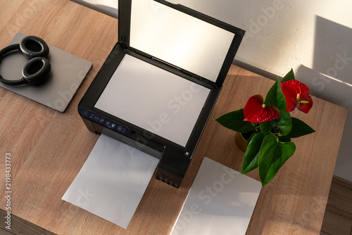 Foto Compact home office setup with open multifunction printer, laptop, headphones, blank paper and flowering plant