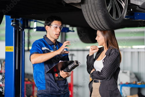 Fotografie Inside a service garage, a customer hears a technician explain battery findings under a lifted EV, with clipboard and work light