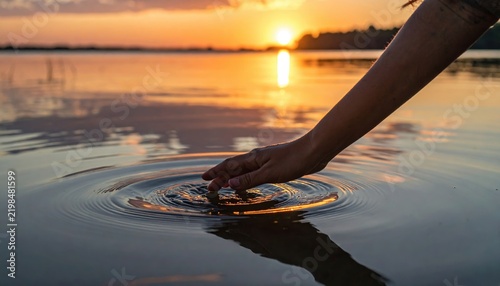 Young female hand touching calm lake water during golden hour sunset creating expanding circular ripples