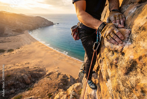 A rock climber ascends a steep cliff overlooking a scenic beach.