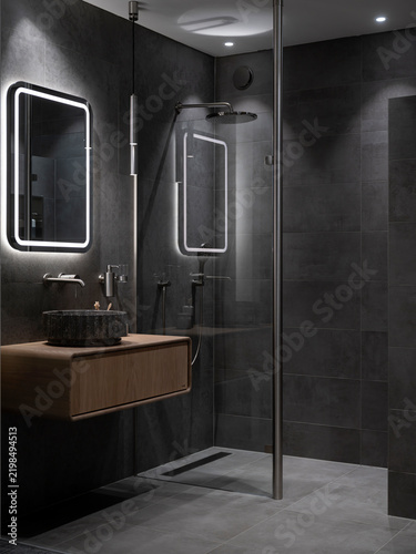 Full view of a dark anthracite bathroom with rain shower, glass partition, stone basin and oak vanity, spotlights cutting through the gloom.