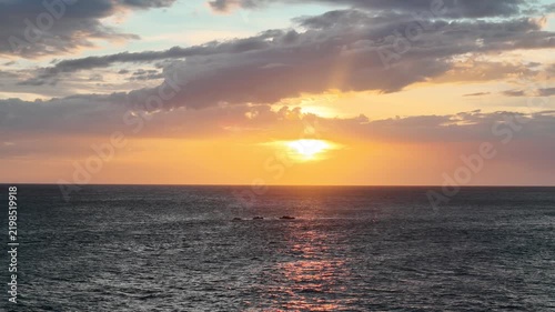 The sun sets over the ocean casting warm colors across the water. Clouds float in the sky as rocks are visible on the horizon.