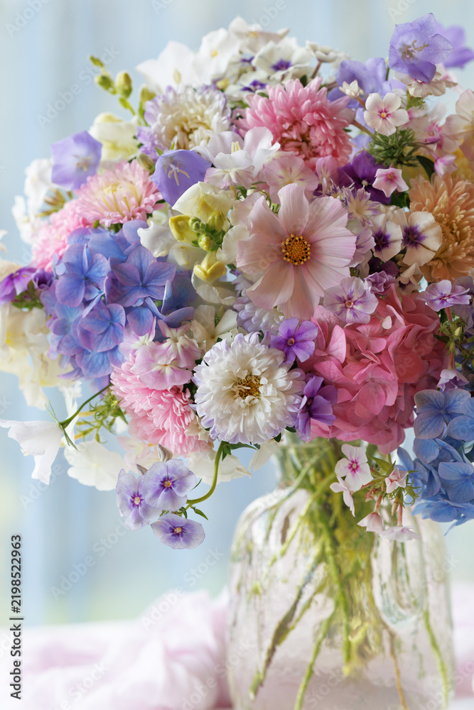 Fototapeta premium Beautiful bouquet in a vase flowers phlox, asters, hydrangeas, and bellflowers on a table, still life closeup set against a window overlooking the garden, blur, selective focus.