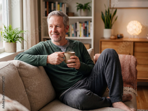 Relaxed middle-aged man holding a warm mug of coffee and smiling while sitting on a living room couch