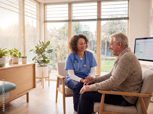 Compassionate female healthcare worker in blue scrubs holding hands and laughing with an elderly male patient in a bright medical clinic