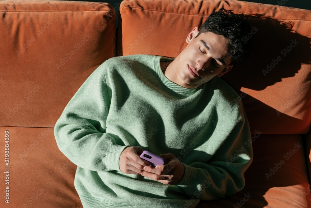 Fototapeta premium Young man asleep on sofa holding smartphone in sunlight for relaxed indoor background and lifestyle imagery