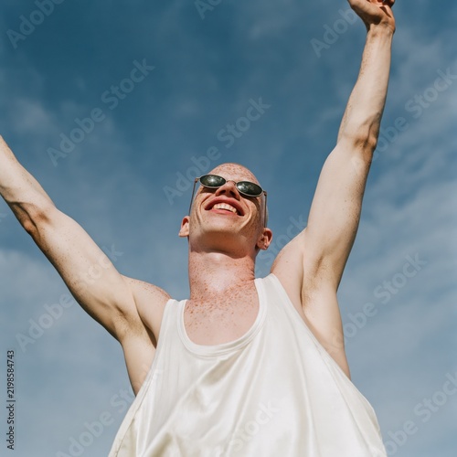 Happy young man stretching arms upward wearing sunglasses under clear blue sky summer outdoor portrait expressing freedom and elation