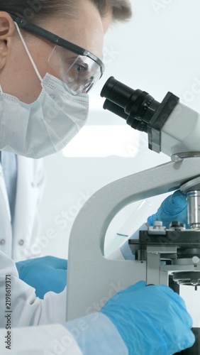 Scientist wearing protective mask, safety glasses, and gloves, examining a slide under a microscope, conducting important medical research and experimental analysis in a laboratory