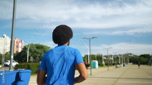 Black amateur athlete with afro hair running along urban park promenade with city buildings in blue sportswear daytime