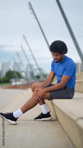 Young Black Latin man with afro hair holding painful knee sitting on steps in urban park with bridge background daytime