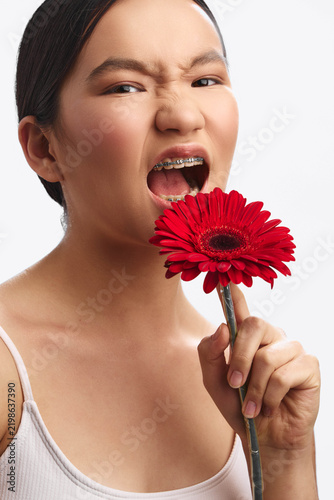 Fotografie Asian woman poses closely with a vibrant red flower held near her open mouth, conveying playful boldness in a bright studio shot with soft, flattering lighting