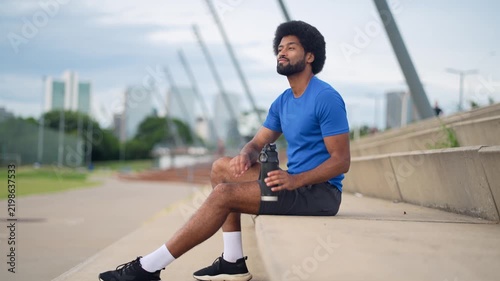 Brazilian amateur athlete with afro hair sitting on steps drinking water from metal bottle with city skyline daytime