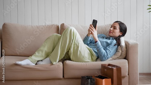 Woman lying on the couch checks her phone, then pulls a vest from a shopping bag and photographs its tag. Relaxed shopper reviewing a receipt or product tag after online shopping on her smartphone