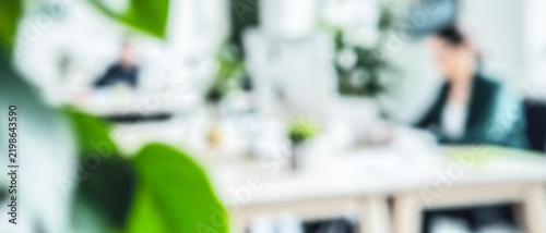 A blurred image of a modern office environment with plants. Two individuals work at desks, focused on their tasks amidst a bright, vibrant backdrop.