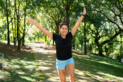 Woman, jumping and celebrating in nature with fitness, joy or achievement outdoor. Athlete or runner with arms up, smart watch and enthusiasm for training success in forest