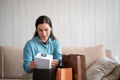 Smiling woman holding a white gift box beside shopping bags on the couch. Female customer checking a purchase after shopping