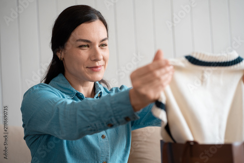 Smiling woman holding up a knitted sweater to inspect it, sitting indoors. Female customer checking a new clothing purchase