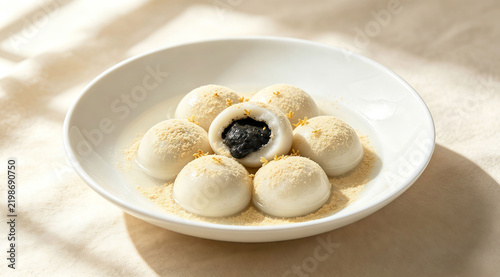 Bitten black sesame tangyuan glutinous rice dumplings sprinkled with soybean powder, served in white ceramic plate with osmanthus petals in light sweet soup