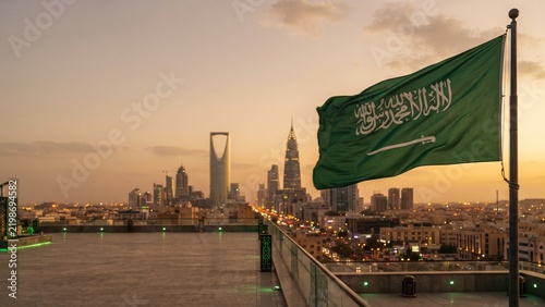 Saudi Arabia national flag waving on flagpole in desert sand dunes at sunset with mountains in background. 