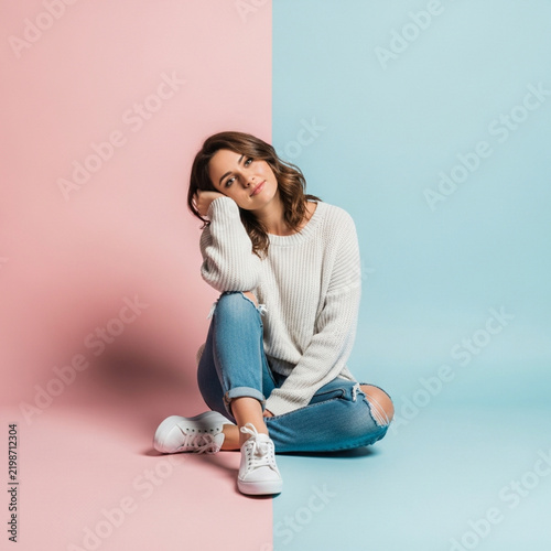 Young Woman Sitting on Floor in Ripped Jeans and Sweater Against Pink and Blue Background