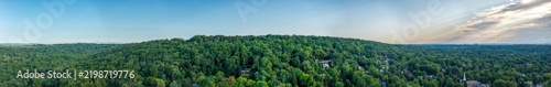 A wide aerial panorama spans green wooded hills and distant treetops beneath a blue sky with a band of pale clouds. The wide composition shows tree covered hills under a partly cloudy sky in daylight.