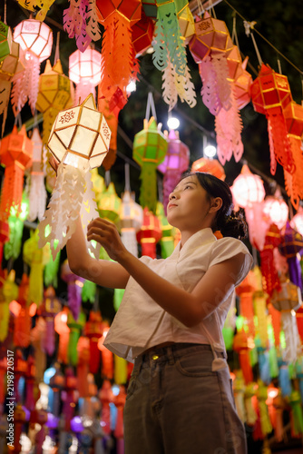 Young Asian woman holding traditional glowing paper Lanna lantern during Yi Peng lantern festival night under colorful hanging lights