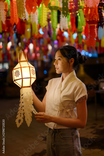 Young Asian woman holding traditional glowing paper Lanna lantern during Yi Peng lantern festival night under colorful hanging lights