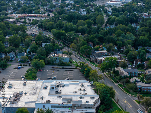 A broad flat building roof, parking spaces and nearby railway tracks contrast with leafy New Jersey surroundings. An aerial view reveals green tree cover beside local buildings under natural daylight.
