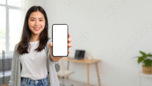 Asian woman holding smartphone with blank white screen, smiling young female showing mobile phone mockup, digital technology in bright modern home office