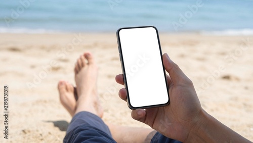 Smartphone mockup with blank white screen held at the beach, person relaxing on sand with mobile phone, digital nomad lifestyle and summer vacation concept