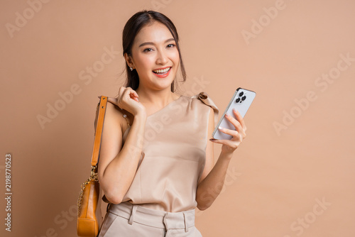 Happy smiling Asian woman holding handbag and smartphone in beige studio, demonstrating fashionable shopping lifestyle, modern mobile banking promotion, consumer satisfaction, and retail elegance.