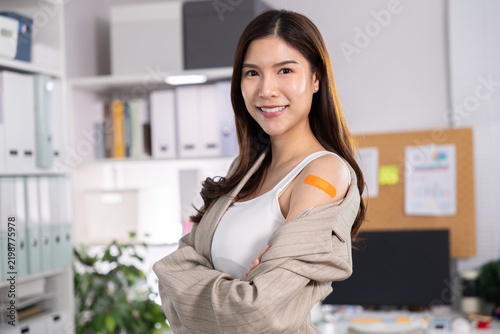 Confident Asian woman with orange adhesive bandage on shoulder standing in modern office, smiling with crossed arms and conveying readiness for workplace wellness and healthcare