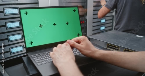 Male System Administrator Uses Laptop With Green Screen Beside Modern Server Cabinets, Creating Flexible Footage For Replacing Display Content With Monitoring Dashboards, Software Interfaces and IT