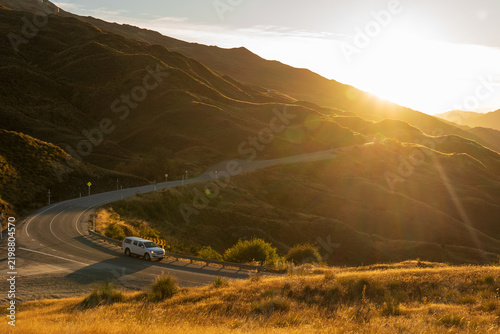 Obraz na plátně Vehicle on a winding mountain road at golden sunset above tussock hills, New Zea